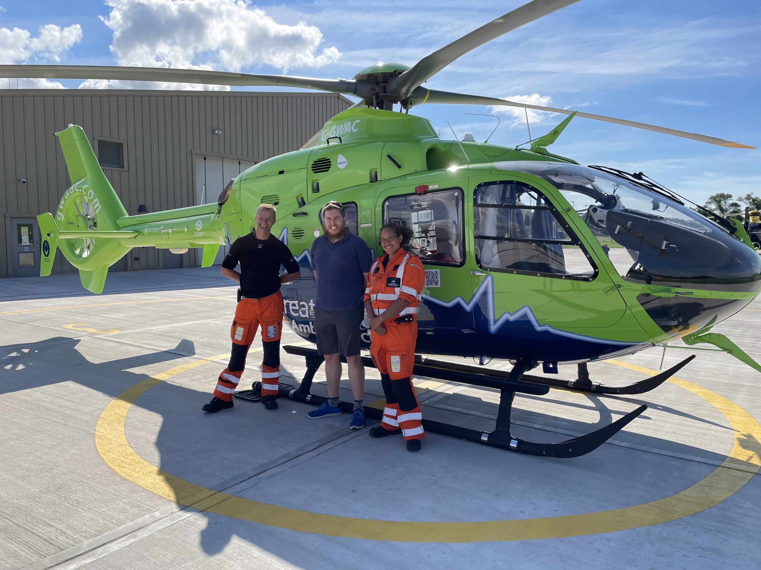 Chris and two paramedics stand in front of the GWAAC lime green helicopter on a bright sunny day.