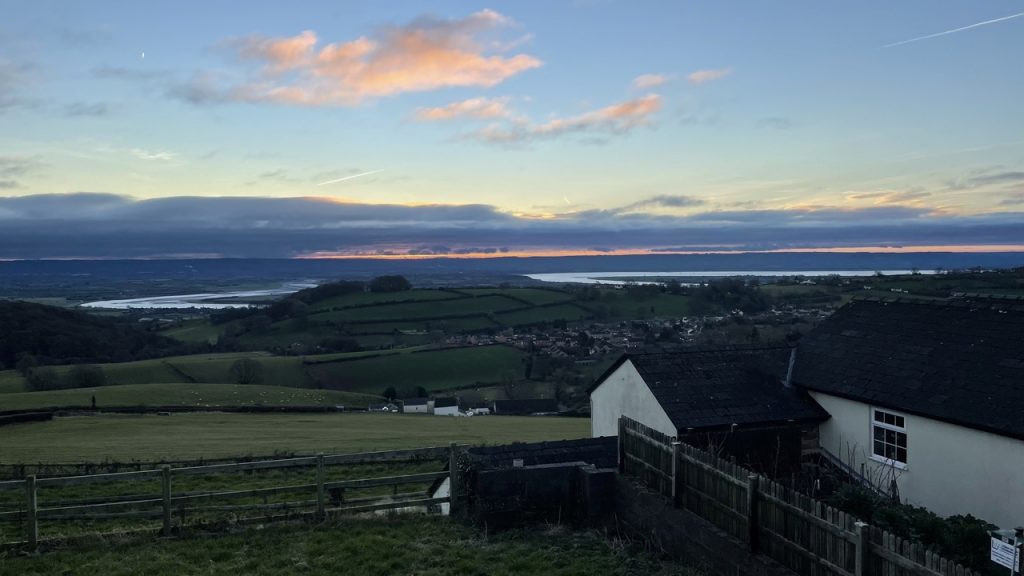 A white gable end and a dry stone wall in the foreground. Green hills fall down into a valley with a band of orange sunrise amonst bands of clouds with blue sky above